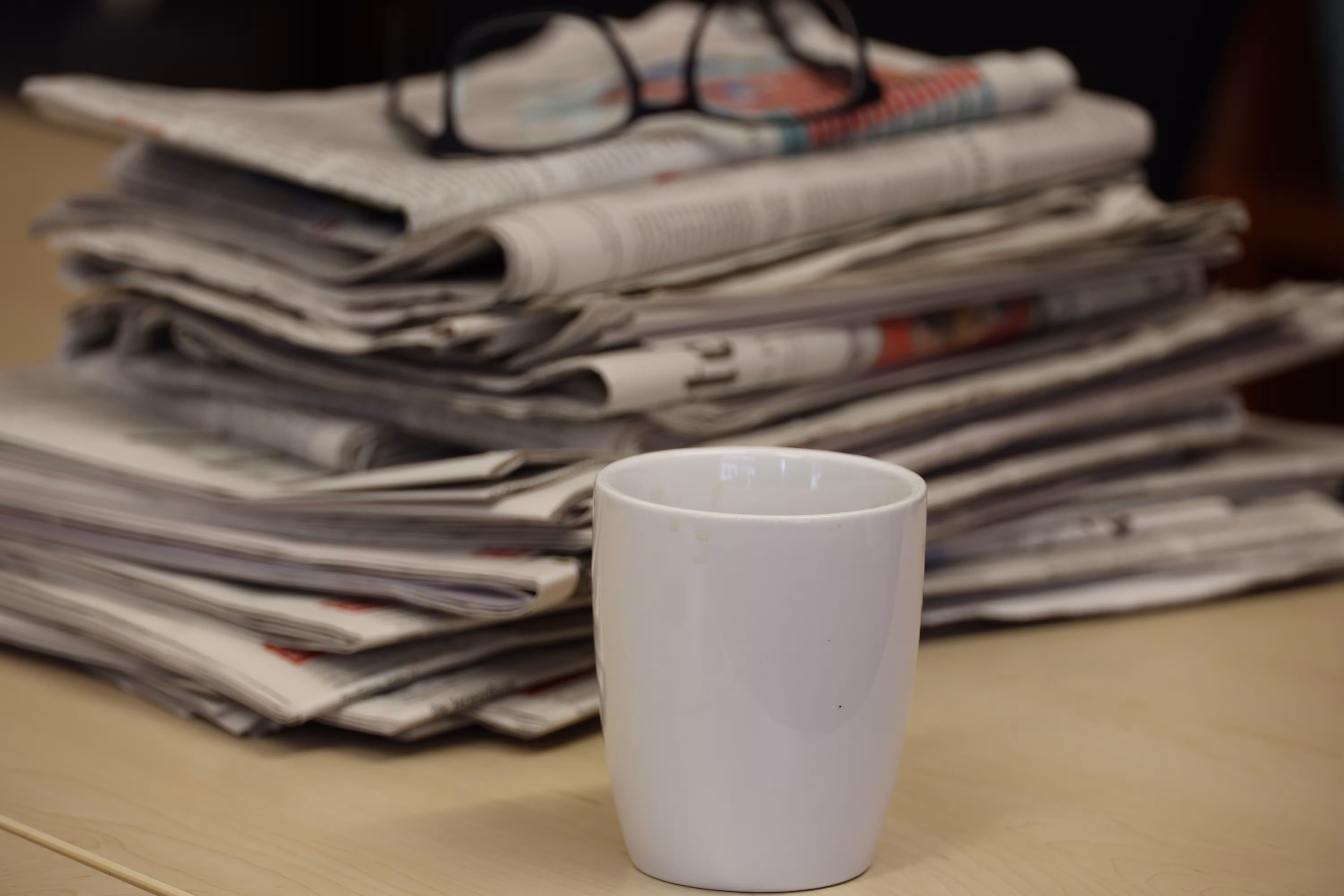 Zeitungsstapel mit abgelegter Brille. Davor steht ein weißer Becher.