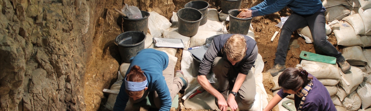 Ausgrabungssituation Hohle Fels, vier Menschen sitzen auf dem sandigen Höhlenboden und arbeiten im Sand. Zwischen ihnen stehen schwarze Eimer. Links von der Arbeitsstelle sind viele weiße Sandsäcke zu sehen, die den restlichen Boden bedecken.