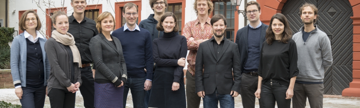 Eine Gruppe von Menschen steht in zwei Reihen vor dem Gebäude der Akademie im Innenhof. Die zwölf Frauen und Männer sind zur Kamera hingewendet und lächeln.