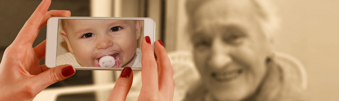 Im Vordergrund sind zwei Hände zu sehen, die ein Smartphone halten. Darauf ist das Bild eines Kleinkinds mit Schnuller zu erkennen. Die Kamera des Smartphones ist auf eine Person im Hintergrund gerichtet, die allerdings nicht mit dem Kleinkind auf dem Bildschirm identisch ist, sondern eine alte Frau mit weißem Haar, die in die Kamera lächelt.