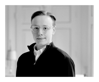 Dreiviertelporträt von Moritz Gaspard in Schwarzweiß. Er trägt eine Brille und schaut direkt zum Betrachter. Die kurzen Haare hat er mit Seitenscheitel zur Seite gekämmt. Über einem weißen Hemd trägt er einen Troyer mit Reißverschluss.