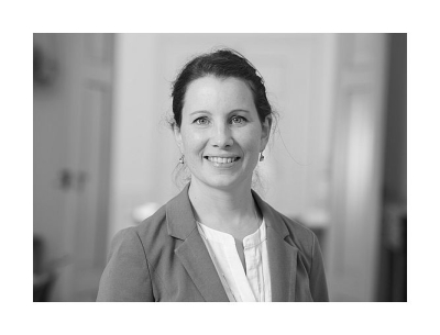 Schwarz-weiß Portrait von Dieta Svoboda-Baas in halbnaher Aufnahme, sie trägt eine weiße Bluse, darüber einen hellen Blazer und lächelt in die Kamera, die Haare sind nach hinten hochgesteckt, dazu trägt sie hängenden Ohrringe mit runden Edelsteinen
