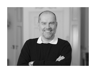 Schwarz-weiß Portrait von Andreas Dafferner in halbnaher Aufnahme, er trägt einen dunkeln Pullover, darunter ragt ein weißer Hemdkragen hervor, die Haare sind kurz rasiert, die Arme vor der Brust verschränkt