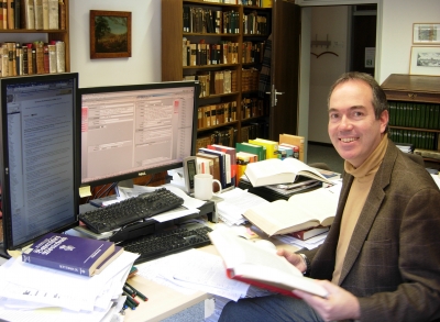Andreas Deutsch an seinem Arbeitsplatz. Er sitzt vor zwei Bildschirmen und einem Schreibtisch voller Bücher. Er hält ein aufgeklapptes Buch in der Hand und hat sich vom Tisch abgewandt, um in die Kamera zu lächeln. Im Hintergrund sind hohe Bücherregale an der Wand des Raumes zu sehen.