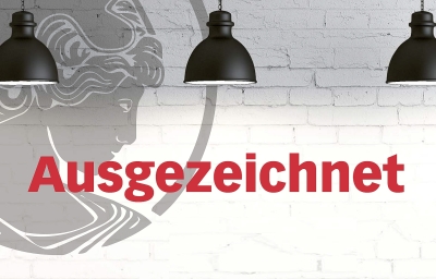 Das Bild zeigt eine weiß gekachelte Wand vor der drei schwarze Lampenschirme hängen. Sie beleuchten das in roter Schrift abgebildete Wort "Ausgezeichnet". In der linken Bildhälfte ist das Logo der Akademie, die Athene im Seitenprofil, zu erkennen, die in grau transparent an die Wand gemalt ist.