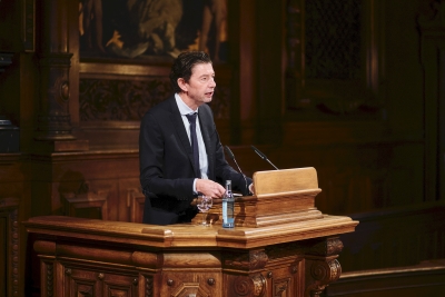 Dreiviertelporträt von Prof. Christian am Pult stehend und sprechend. Er hat dunkle lockige Haare und trägt einen Anzug mit weißem Hemd und Krawatte.