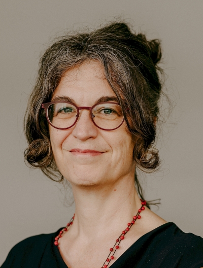 Portrait von Claudia Diehl in Nahaufnahme. Sie lächelt in die Kamera, trägt die Haare hochgesteckt und eine rotgerahmte Brille. Dazu trägt sie eine schwarze Bluse und eine Kette mit roten Perlen.