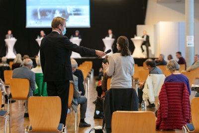 Raumaufnahme eines Vortragssaals, im Vordergrund Stuhlreihen, zwischen denen Herr Brock einer Teilnehmerin das Mikrofon für eine Publikumsfrage hinhält, er steht seitlich zu den Betrachtenden und trägt einen medizinischen Mundschutz, die Teilnehmerin steht mit dem Rücken zur Kamera, im Hintergrund eine Bühne mit Stehtischen für eine Podiumsdiskussion