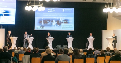 Raumaufnahme eines Vortragssaals, eine PowerPoint wird an eine schwarze Wand gestrahlt, vor der fünf zwei bezogenen Stehtische für eine Podiumsdiskussion stehen. An jedem Tisch steht eine Person, im Vordergrund des Bildes ist das Publikum mit dem Rücken zur Kamera gedreht zu sehen