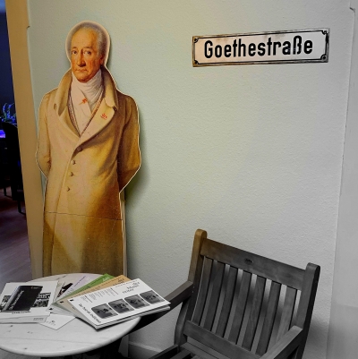 Im Vordergund steht ein runder Beistelltisch mit einem hölzernen Stuhl. An die Wand gelehnt steht hinter dem Tisch ein Pappaufsteller von Goethe. Rechts daneben hängt ein Straßenschild mit der Aufschrift "Goethestraße".