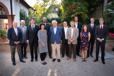 Gruppenbild im Garten der HAdW, im Hintergrund sind grüne Pflanzen zu sehen, davor stehen 12 Personen in zwei Reihen zusammen, sie tragen feierliche Kleidung und lächeln in die Kamera, die 10 Männer tragen dunkle Anzüge, eine Frau in der ersten Reihe links ein graues Kleid, eine Frau in der Resten Reihe rechts ein blaues Kleid mit rotem Blumenmuster