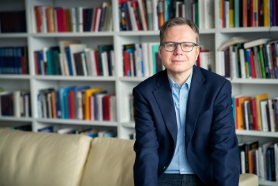 Das Bild zeigt Andreas Reckwitz im Anzug mit offenem Jacket auf der Lehne eines Sofas sitzend. Im Hintergrund ist eine Bücherwand zu sehen.