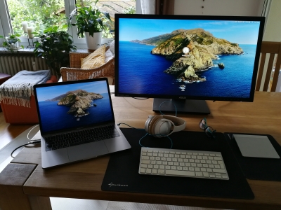 Ein Bildschirm und ein Laptop mit einer Felsinsel als Bildschirmschoner stehen auf einem hölzernen Tisch. Dahinter ist eine Wohnzimmereinrichtung zu sehen.