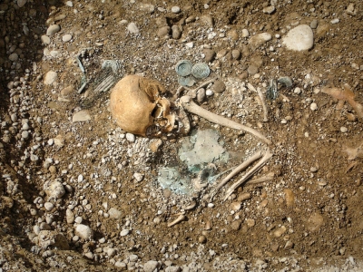 Bild eines aus dem Sandboden ragenden Skeletts. Der Kopf, die Schulter sowie ein Arm sind zu sehen. Neben dem Skelett liegen einige Schmuckstücke.