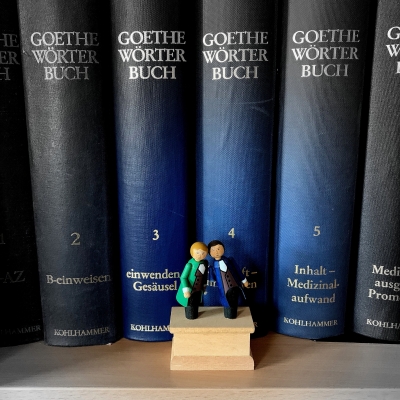 Aufnahme von nebeneinander aufgereihten Büchern mit dunkelblauem Einband, es sind Werke der Reihe "Goethe Wörterbuch" wie dem silbernen Aufdruck auf den Buchrücken zu entnehmen ist. Davor steht eine Miniaturfigur von zwei im klassischen Stil der Zeit Goethes gekleideten Männern, deren Köpfe gegeneinander gelehnt sind.