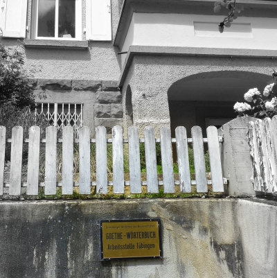 Blick auf das Gebäude der Arbeitsstelle Goethe-Wörterbuch in der Frischlinstraße 7 in Tübingen. Im Vordergrund sind eine graue Mauer und ein weißer Holzzaun vor einem weiß verputzten Haus zu sehen. Von dem Haus ist nur ein kleiner Ausschnitt des Eingangs sowie eines Fensters zu sehen. Auf der Mauer im Vordergrund ist ein goldenes Schild mit schwarzer Aufschrift angebracht, auf dem steht. "Goethe Wörterbuch. Arbeitsstelle Tübingen"