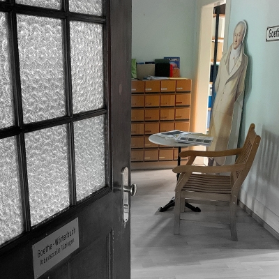 Eingang zu den Räumlichkeiten der Goethe-Wörterbuchs in Tübingen. Links eine geöffnete braune Holztür mit Milchglasscheiben, dahinter öffnet sich ein Raum mit einer Kommode an der hinteren Wand. Davor steht ein runder Beistelltisch mit einem hölzernen Stuhl. An die Wand gelehnt steht hinter dem Tisch ein Pappaufsteller von Goethe. Zwischen der Kommode und der Figur liegt eine Türöffnung, die in einen weiteren Raum führt.