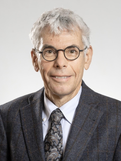 Brust-Porträt von Stefan Maul. Er trägt eine runde Brille hat kurze graue Haare und lächelt den Betrachter/die Betrachterin an. Er trägt ein Hemd, Krawatte und eine Anzugsjacke.