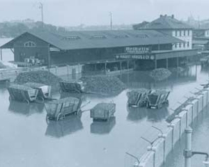 Schwarz-weiß Foto des Elbehochwassers im Januar 1932: Die Elbe tritt in der Nähe von Dresden über ihre Ufer (Bundesarchiv, Bild 102-12896, anonymes undatiertes Foto); zu sehen sind ein längliches Gebäude und einige Kutschwagen, deren Räder im Wasser stehen.