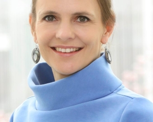 Portrait in halbnaher Aufnahme von Lena Maier-Hein. Sie trägt einen hellblauen Pullvoer und lächelt frontal in die Kamera. Die Haare trägt sie nach hinten hochgesteckt, dazu große silberne Ohrringe.