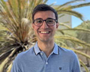 Portrait eines braunhaarige Mannes mit Brille vor einer Palme. Er trägt ein hellblaues Polo-Hemd.