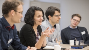 Das Foto zeigt eine Aufnahme von vier Personen an einem Tisch von der Seite aufgenommen, links ein Mann, der zu der Frau zu seiner Linken herübersieht, die Hände auf dem Tisch ruhend. Die Frau spricht, den Blick geradeaus zur rechten Bildseite gerichtet und hat die Ellbogen auf der Tischplatte abgestützt, die Hände vor der Brust geöffnet. Links von ihr ein Mann, der in die gleiche Richtung wie sie schaut. Ganz links am rechten Bildrand, ein dritter Mann, dieser schaut zu der sprechenden Frau.