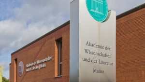 Aufnahme des Gebäudes der Akademie der Wissenschaften Mainz. Vor dem roten Backsteinbau mit Flachdach, an dessen Wand das Wappen mit dem Namen der Akademie angebracht ist, steht eine metallische, rechteckige Installation, in die der Name der "Akademie der Wissenschaften und der Literatur Mainz" eingraviert ist. Oben hat die Installation eine halbrunde Auskerbung, in die das runde Wappen der Akademie in türkis eingelassen ist.