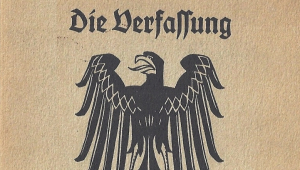 Das Bild zeigt das Deckblatt der Verfassung von Weimar. Auf beigen Untergrund ein schwarzer Adler zu sehen, über dem die Worte "Die Verfassung" stehen. Unterhalb des Adlers steht "des Deutschen Reiches". 
