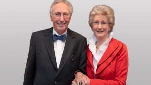 Das Ehepaar Josephine und Hans-Werner Hector in der Halbtotalen. Hans-Werner Hector trägt einen schwarzen Anzug mit weißem Hemd und blau-grauer Fliege. Josephine Hector trägt einen roten Blazer und eine weiße Bluse und hat sich am Arm ihres Mann untergehakt.