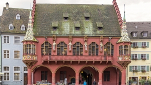 Rote Fassade des Historischen Kaufhauses am Münsterplatz in Freiburg. Im Erdgeschoss überdachte Laube mit vier sichtbaren Arkaden; darüber zieht sich ein langer Balkon, der unter fünf spätgotischen Vorhangbogenfenstern verläuft, die an den Ecken jeweils mit einem Erker abschließen. 
