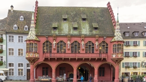 Rote Fassade des Historischen Kaufhauses am Münsterplatz in Freiburg. Im Erdgeschoss überdachte Laube mit vier sichtbaren Arkaden; darüber zieht sich ein langer Balkon, der unter fünf spätgotischen Vorhangbogenfenstern verläuft, die an den Ecken jeweils mit einem Erker abschließen.
