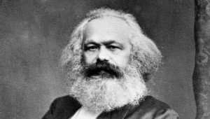 Schwarzweiß-Foto von Karl Marx. Er sitzt in einem Stuhl und blickt den Betrachter frontal an. Seine rechte Hand steckt er dabei unter das Revers seines Gehrocks, der linke Arm ruht auf der Stuhllehne.  