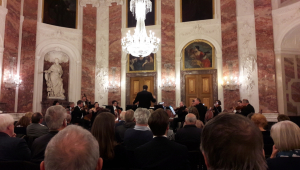 Im Rittersaal im Mannheimer Schloss ist über die Köpfe des Publikums hinweg ein Musikensemble zu sehen. Dieses sitzt vor zwei großen hölzernen Türen. Die Wände des Saals sind in weißem und rotem Marmor gestaltet und werden von Wandgemälden und einer Skulptur geziert.