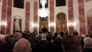 Im Rittersaal im Mannheimer Schloss ist über die Köpfe des Publikums hinweg ein Musikensemble zu sehen. Dieses sitzt vor zwei großen hölzernen Türen. Die Wände des Saals sind in weißem und rotem Marmor gestaltet und werden von Wandgemälden und einer Skulptur geziert.