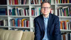 Das Bild zeigt Andreas Reckwitz im Anzug mit offenem Jacket auf der Lehne eines Sofas sitzend. Im Hintergrund ist eine Bücherwand zu sehen. 
