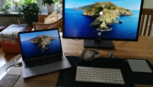 Ein Bildschirm und ein Laptop mit einer Felsinsel als Bildschirmschoner stehen auf einem hölzernen Tisch. Dahinter ist eine Wohnzimmereinrichtung zu sehen.