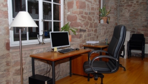 Arbeitsplatz in der Bibliothek der HAdW: Ein Schreibtisch mit Comupter und Monitor steht vor einem weiß gerahmten Fenster. Die Wand ist gemauert mit verschieden großen roten Steinen und grauen Fugen. Vor dem Schreibtisch steht ein lederner Schreibtischstuhl.