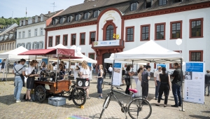 Im Vordergrund wird Kaffee ausgeschenkt und Menschen informieren sich an verschiedenen Ständen über Projekte des Wissenschaftlichen Nachwuchses. Im Hintergrund ist das Akademiegebäude.  