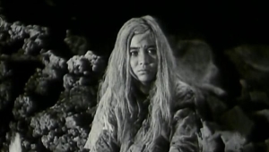Still aus dem in schwarzweiß gedrehten chinesischen Film „Bai Mao Nü” von 1950, der das namengebende weißhaarige Mädchen sitzend in einer Höhle zeigt.