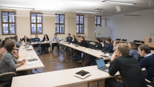 Szene aus einer Veranstaltung der Jungen Akademie im Hörsaal der Akademie der Wissenschaften. Die Teilnehmer:innen sitzen an Tischen, die in einem Rechteck angeordnet sind. Gegenüber vom Betrachter, auf der linken Seite gestikuliert einer der Teilnehmer. Die anderen Teilnehmer:innen folgen seinen Ausführungen.