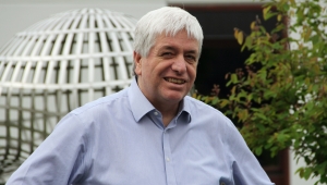 Foto von Gerhard Huisken, der den Betrachter anlächelnd bis zur Brust dargestellt ist. Das Foto zeigt ihn im Hemd mit weißen kurzen Haaren, die nach vorne gekämmt in die Stirn fallen. Im Hintergrund ist ein grüner Baum, sowie ein Pflanzengitter zu sehen. Beides steht vor einem Gebäude, dessen offenen Eingang man sieht.