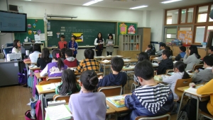 Schulklasse von hinten mit Blick zur Lehrerin und zwei Schülerinnen, die vorne vor der Tafel stehen.