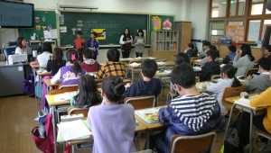 Schulklasse von hinten mit Blick zur Lehrerin und zwei Schülerinnen, die vorne vor der Tafel stehen.
